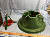 Green plastic Christmas tree stand with five metal screws, red tips, round base for real trees, alongside smaller red and green metal stand for artificial trees. A measuring stick lies in front.