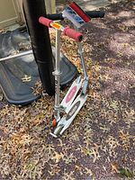 Pink grip Razor scooter standing upright on a brown leafy outdoor surface, frame, wheels, and handlebars visible with some dirt.