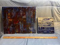 Two unframed abstract paintings placed side by side on a white cloth background with a measuring ruler below them showing sizes approximately 15-16 inches wide for larger canvas and about 8-9 inches for smaller canvas.