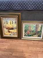 Two framed landscape paintings resting on floor, one with birch trees by water and another with autumn forest path