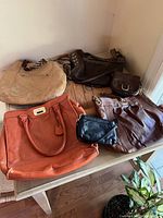 Seven leather handbags arranged on wooden surface, showing various colors and styles including tan, brown, black and rust-orange perforated bag.