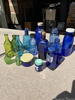 Photo of 16 vintage glass bottles in various shades of blue and green, arranged in several rows on flat surface in sunlight.