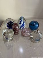 Front view of 7 glass paperweights including apples, spherical and egg shapes showing colors and design variety.