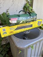 Packaged Coleman 4 person tent in green carry bag with Coleman logo, and boxed Byer Allagash Plus cot on top of outdoor air conditioning unit surrounded by ivy.