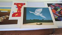 Four large art prints spread flat on table: Salvador Dali's 'The Persistence of Memory' with melting clocks, Magritte's 'The Return' showing a bird in flight, Vasarely's 'Tridem-K' geometric color pattern, and a red print titled 'Out of Sight' with faces and birds.