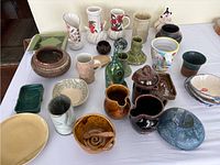 Photo of entire assortment of handmade pottery items displayed on a white cloth, showing variety of shapes, sizes, and colors including vases, pitchers, bowls, mugs, and jars.