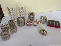 Seven chrome candle holders with animal print and striped cutout designs, two ceramic containers with teddy bear lids, unopened box of 3 silver wine stoppers with grape design, and small Christmas tree decorations in boxes with tiny balls visible.