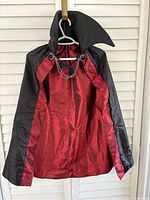 Front view of the cape hanging showing the black velvet exterior, red satin lining, upright collar, and chain closure.