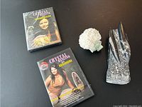 Two DVD cases and two mineral specimens shown on a black surface: Crystal Healing DVDs 'Intermediate' and 'Beginners,' a white spherical fibrous crystal with green cubic crystals, and a tall dark Orthoceras fossil.