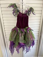 Purple velvet fairy dress with green and purple chiffon layered skirt hanging on hanger in front of shuttered window