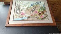 Framed needlepoint artwork depicting a nature landscape with mountain, waterfall, trees, flowers, bridge, and watermill.