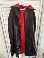 Front view of a full-length Dracula-style cape, black on the outside with a red lining and a collar, hanging on a white hanger against a white louvered backdrop.