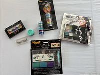Photo of Halloween makeup items including striped false eyelashes, blue face paint, rainbow stack face paint, 'Haunting' eye shadow palette, and Spirit Peacock cosmetic kit.