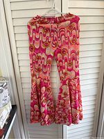 Full view of hippie flared bell bottom pants hanging on a hanger against a white shutter background showing their overall psychedelic pattern and shape.