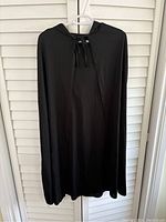 Front view of black lightweight hooded cape hanging on a hanger, showing tie closure at neckline and smooth fabric