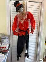 Full view of skeleton scarecrow wearing red shirt and black pants with a black top hat in front of white folding doors.