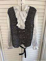Front view of men's black and white Victorian style frilled shirt with floral vest pattern and silver buttons, hanging on white hanger.