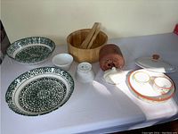 Multiple serving dishes including green floral pattern oval plates, wooden salad bowl with utensils, white ceramic small containers and garlic roaster box.