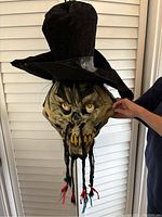 Scary ghoul mask with top hat displayed against white shutter background, showing detailed face, large eyes, yellow teeth, and hanging braids.