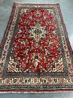 Full view of rectangular red field rug with floral and vine border laid on concrete floor
