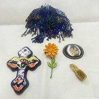 Five assorted brooches displayed on white background including a blue beaded fringe brooch, a colorful ceramic cross, an orange flower, oval black brooch with leaves, and gold-tone pineapple.
