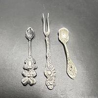 Three antique sterling silver mini flatware pieces: two spoons and one fork, displayed flat with detail of ornate handles and size reference.