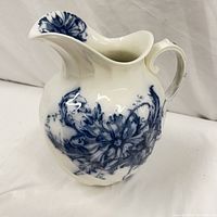 Photo showing side view of white ironstone pitcher with blue floral transferware design, glossy ceramic surface, curved handle and flared spout.