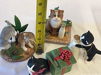 Four Lenox cat figurines placed side by side with measuring tape showing heights: one black and white cat near 4 inches, one orange tabby cat, pair of gray cats on leafy base, and green gift box with bow.