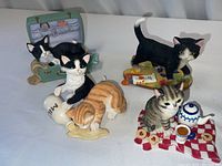 Photo showing all four Lenox cat figurines arranged on white background: cat in jewelry box, cat with milk bottle, orange tabby with spilled milk, cat near tea set.
