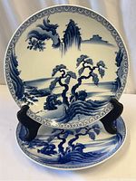 Two Japanese Imari plates displayed with one propped up on stand, showing blue and white painted landscape design with stylized trees, water, and mountains.
