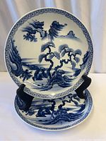 Two Japanese porcelain Imari plates displayed on stands showing the full front blue and white painted landscape design with trees, mountains, and water, including decorative border pattern.