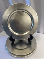 Front view of one metal charger plate on black plate stand, showing matte pewter-like textured surface and round shape with recessed center and raised rim.