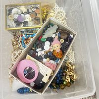 Plastic container with assortment of vintage costume jewelry and buttons including beaded necklaces, plastic buckle, and collectible charms.