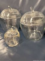 Front angle showing three clear glass apple-shaped jars with matching lids and stem handles on a black background.