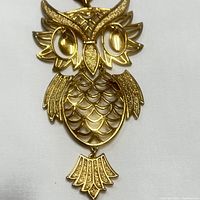 Close-up photo showing detailed textured goldtone owl pendant with articulated wings, scalloped body, detailed eyes, and movable tail piece.
