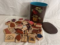 Photo showing 23 vintage wooden rubber stamps with different Arts & Crafts style designs, positioned around a brown container hand-painted with red and yellow apples and green leaves. Another brown lid is also visible.