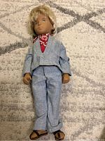 Full body shot of the blonde Sasha Gregor doll dressed in denim outfit with red bandana and sandals, laying on a patterned carpet.