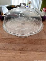 Front view of clear glass cake stand with domed lid on wooden surface showing floral etched pattern and round knob handle.