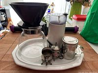 Overview of coffee station setup with silver sugar and creamer set, shaker, Braun coffee grinder, glass pour over coffee pot with black filter holder, on a white Pfaltzgraff platter.