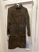 Full view of brown faux suede trench coat hanging on a hanger against white shutters.