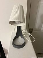 Front view of the IKEA table lamp showing white pleated shade, black oval base, and attached power cord with switch.
