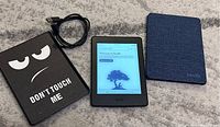Photo showing Kindle Paperwhite 7th gen e-reader powered on with 'Welcome to Kindle' screen, placed on carpet next to two protective cases and a charging cord.