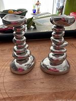 Front view of two silver-tone Michael Aram candle holders shaped like stacked river rocks on wooden surface with background items visible.