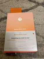 Peach-orange fitness journal with branding 'The Badass Body Goals' and text about booty shaping and resistance training journey.