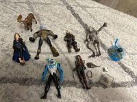 Nine assorted action figures and toys arranged on a floor including humanoid and fantasy creatures, small dinosaur figures, and toy accessories.