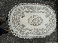 Oval white carpet featuring gold and green embroidery with tasselled edges laid on a dark textured ground surface.