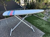 Ironing board outdoors showing padded cover with blue and red stripes and metal legs.