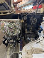 Photo showing two planters side by side. The black oriental metal planter with curved legs and painted decorated figures is on the right. The round ceramic floral planter on a black carved stand is on the left.