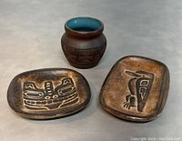 Photo of two rectangular handmade clay dishes with Native Canadian designs (raven and Haida bear) alongside a small signed pot with carved geometric patterns and turquoise interior.