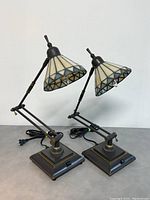 Pair of Tiffany style stained glass swing arm desk lamps with geometric stained glass shade and metal swing arm base with on/off switch on base.